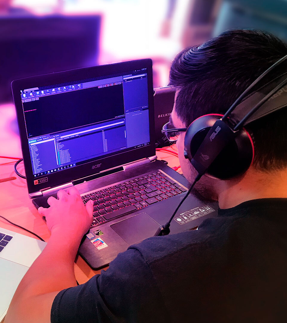 Photo of a young man sitting in front of a laptop computer. He is looking at the screen concentrated while typing source code. He wears large black headphones. The image evokes a young web developer working on coding a website.